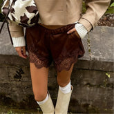 Person wearing brown lace shorts with a beige jacket and white boots, standing against a stone wall.